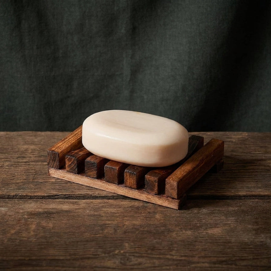 A cream-colored oval bar of soap resting on a dark brown wooden soap dish, set on a rustic dark wood table against a dark fabric backdrop.