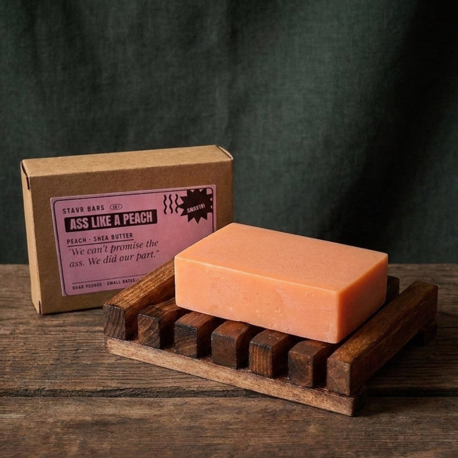A rectangular peach-colored soap bar resting on a dark wooden soap dish, with its kraft paper packaging box behind it on a rustic wooden table against a dark green fabric backdrop.