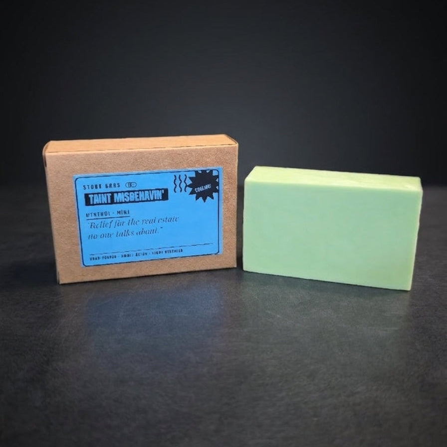 A pale mint green soap bar next to its brown cardboard packaging box with a blue label, set against a dark black background.