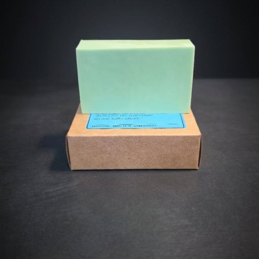 A pale mint green rectangular bar of soap standing upright on top of a brown kraft paper box with a blue label, set against a dark gray background.