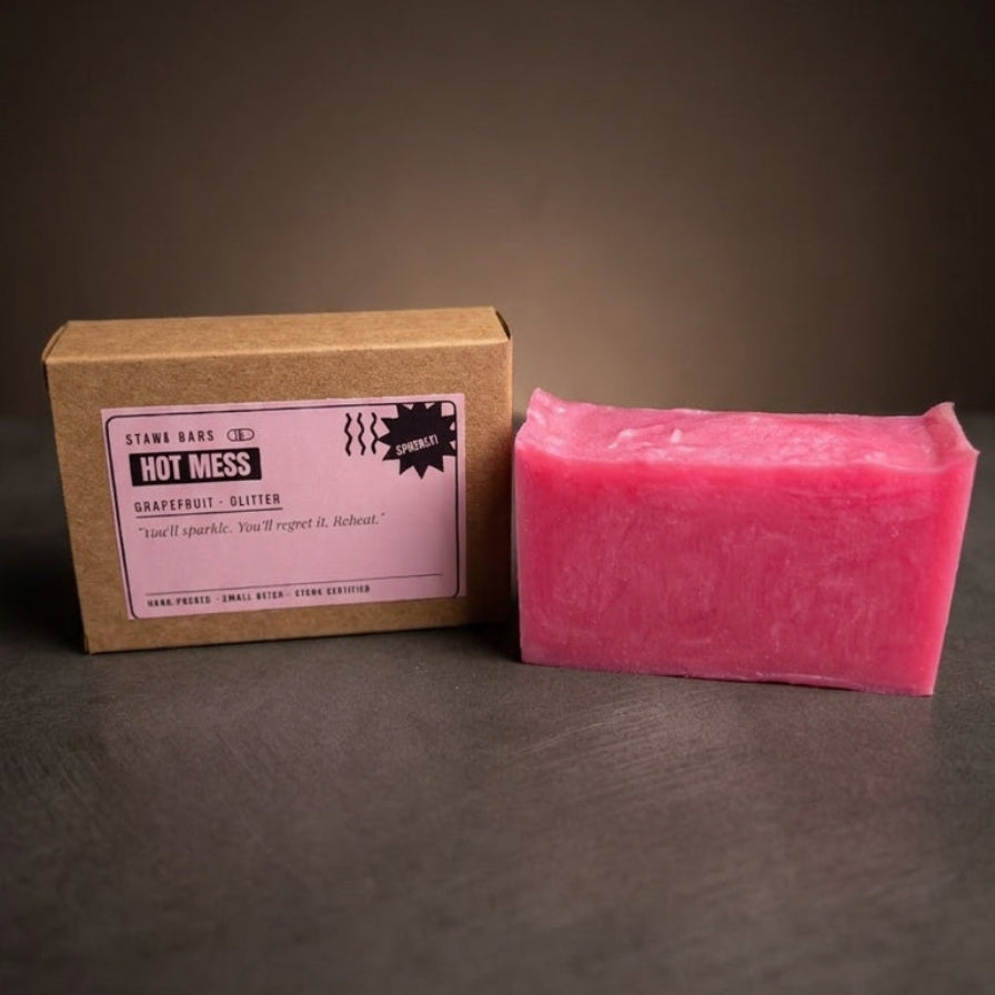 A pink rectangular bar of soap stands next to its brown kraft paper box with a pink label on a dark gray surface against a dark brown background.