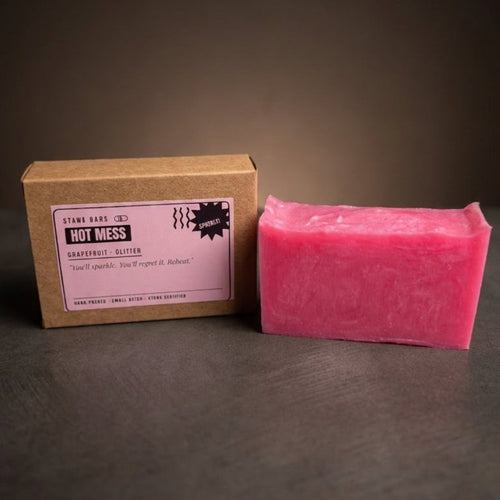 A pink rectangular bar of soap stands next to its brown kraft paper box with a pink label on a dark gray surface against a dark brown background.
