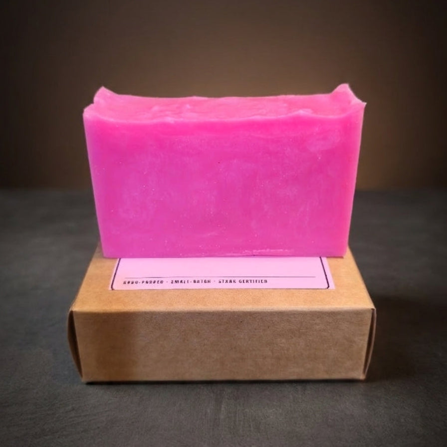 A bright pink rectangular bar of soap standing upright on top of a brown kraft paper box against a dark gradient background.