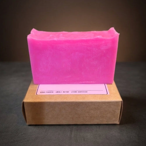 A bright pink rectangular bar of soap standing upright on top of a brown kraft paper box against a dark gradient background.