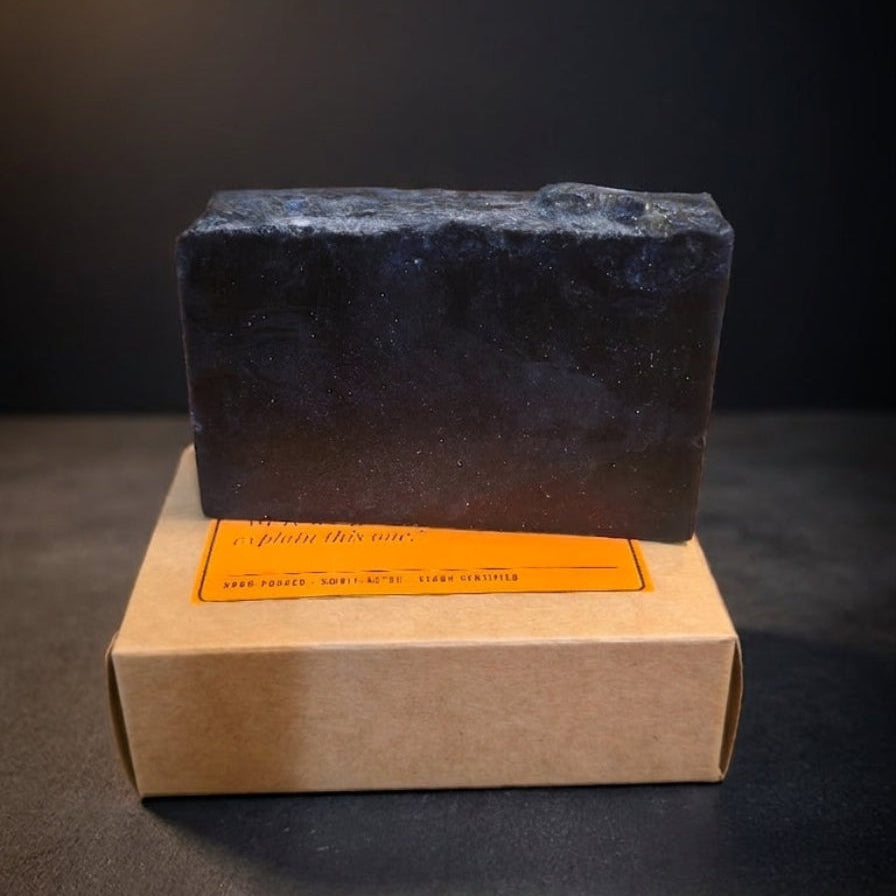 A dark charcoal-colored rectangular bar of soap resting on top of a brown kraft paper box with an orange label, set against a dark studio background.