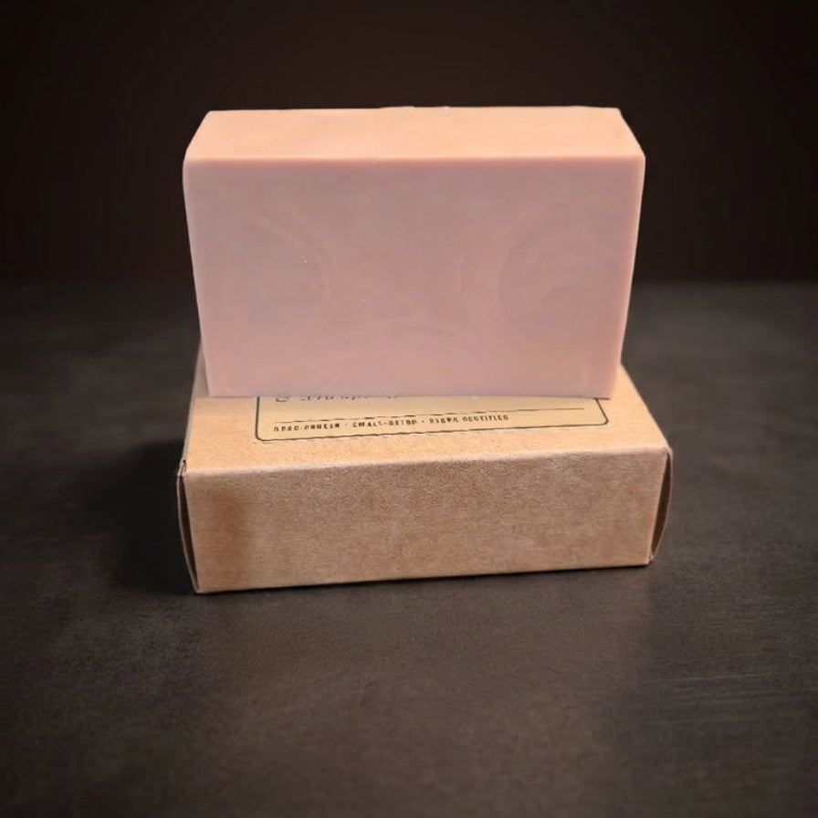A pale pink rectangular bar of soap standing upright on top of a plain kraft paper box against a dark background.