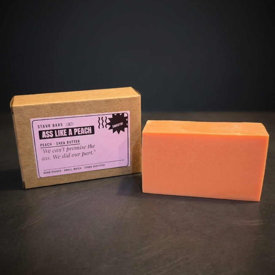 An orange rectangular bar of soap stands next to its kraft paper packaging box on a dark surface against a dark background. The box has a white label that reads 'Piss Like & Peach' and shows the brand 'The Real Soap Co.