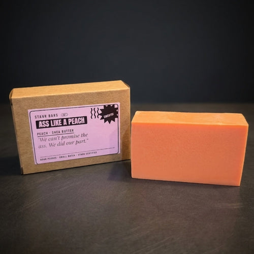 An orange rectangular bar of soap stands next to its kraft paper packaging box on a dark surface against a dark background. The box has a white label that reads 'Piss Like & Peach' and shows the brand 'The Real Soap Co.