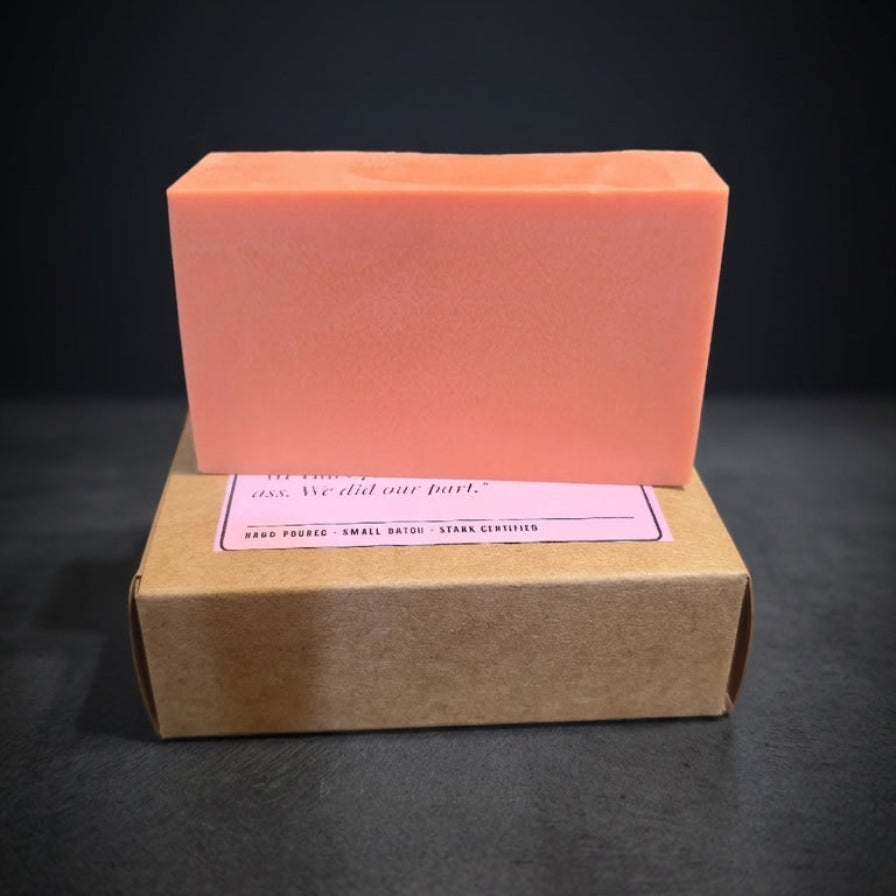 A rectangular peach-colored soap bar stacked on top of a brown kraft paper box with a pink label, set against a dark black background.