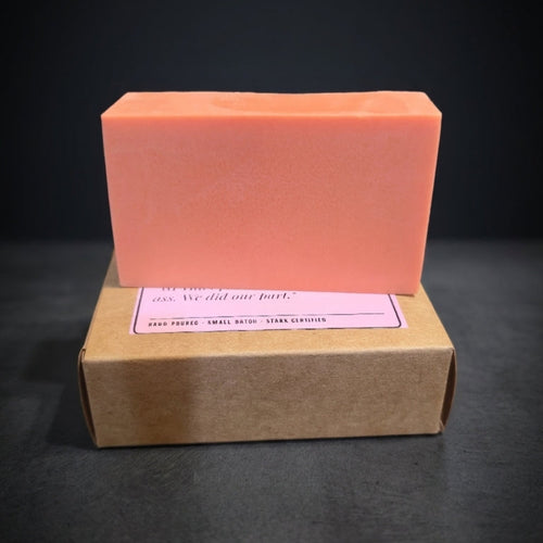 A rectangular peach-colored soap bar stacked on top of a brown kraft paper box with a pink label, set against a dark black background.