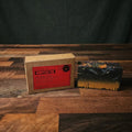 A handmade rectangular bar of soap with a black, gray, and orange marbled pattern sits next to its kraft paper packaging box with a red label, displayed on a rustic wooden surface.