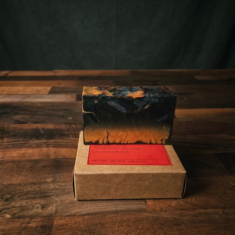 A rectangular bar of soap with a black and orange marbled pattern sits on top of a brown kraft paper box with a red label, placed on a dark wooden table against a dark backdrop.