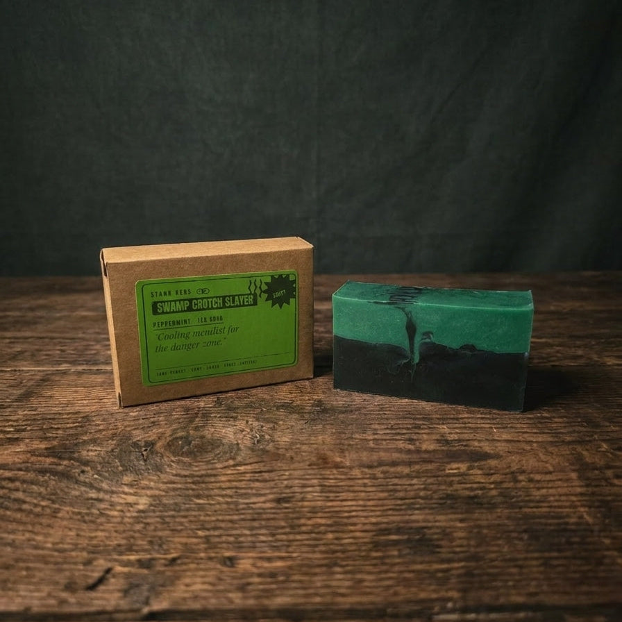 A handmade rectangular bar of soap in green and black sits on a rustic wooden table next to its brown kraft paper packaging box with a green label.