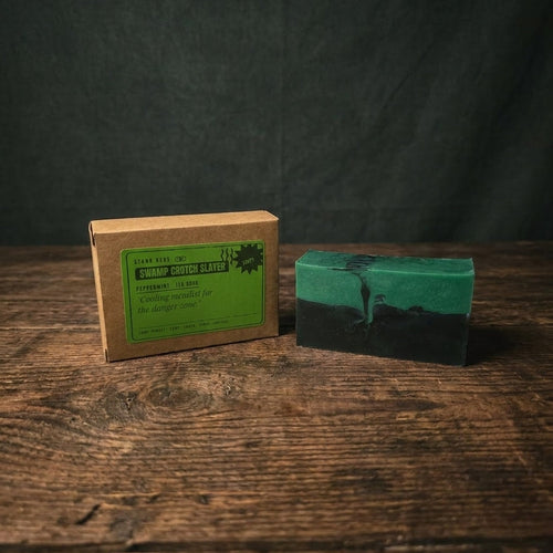 A handmade rectangular bar of soap in green and black sits on a rustic wooden table next to its brown kraft paper packaging box with a green label.