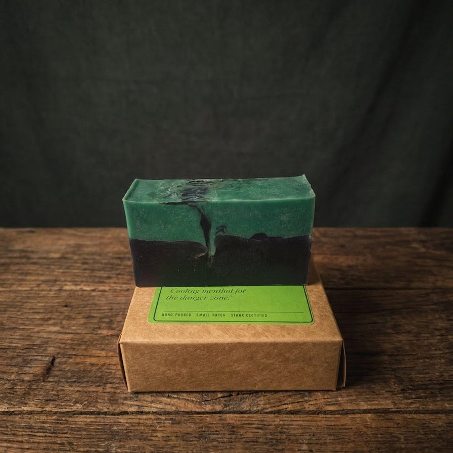 A rectangular bar of soap with a green and black marbled pattern sits on top of a small kraft paper box with a green label, placed on a rustic wooden surface against a dark backdrop.