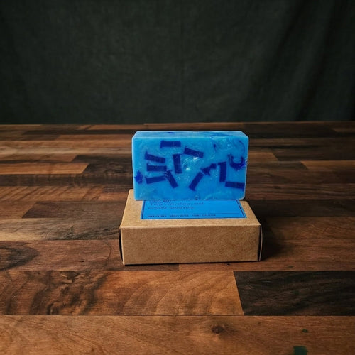 A blue rectangular bar of soap with dark blue abstract markings, displayed on top of a brown kraft paper box on a wooden tabletop against a dark backdrop.