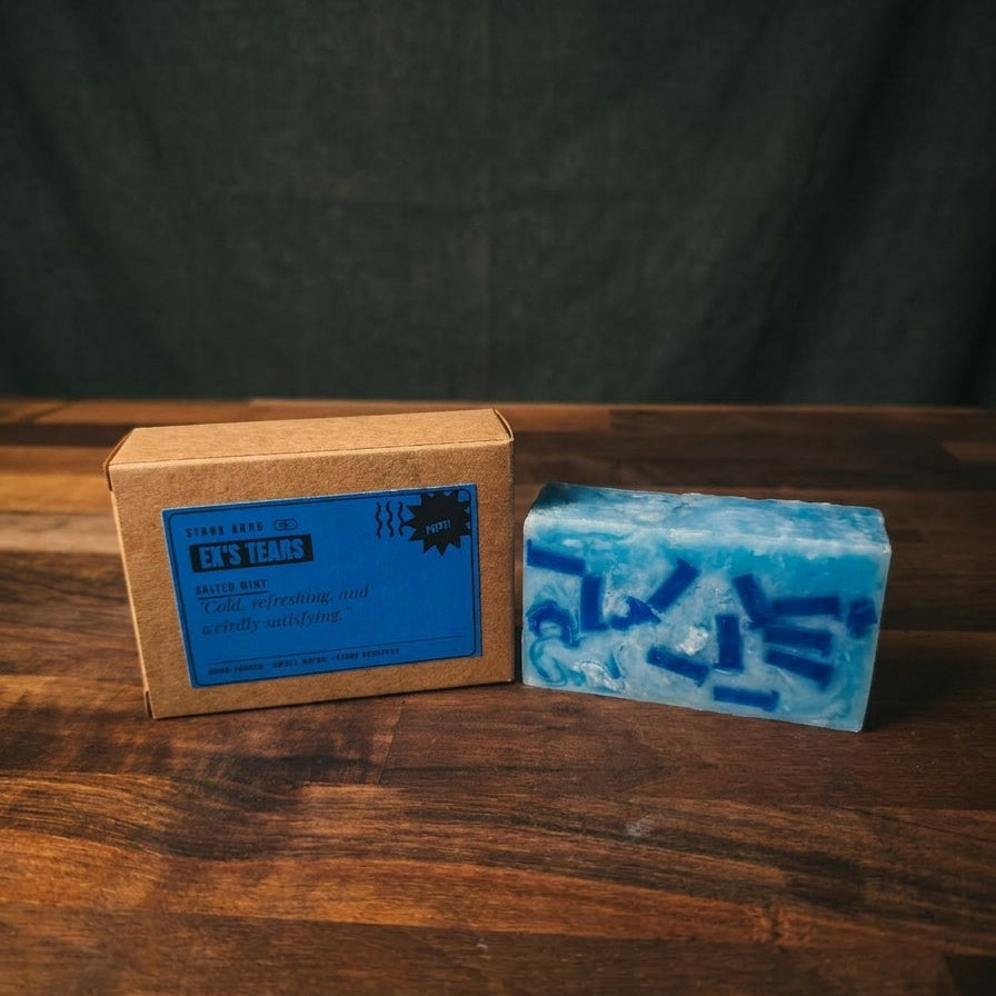 A handmade blue soap bar with dark blue inclusions sits next to its brown kraft paper packaging box with a blue label, set on a dark wooden table against a dark fabric backdrop.